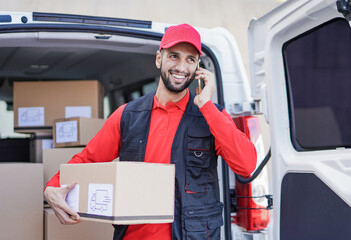 Young caucasian courier man calling clients with smartphone while holding delivery box out of van
