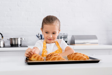 Smiling kid in apron touching homemade croissant on blurred foreground in kitchen