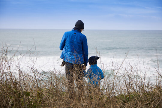 Young dad with glasses and cap is seen from behind. Carries a baby and speaks to his son. View through the grasses. In the background is the sea. Day. Normal perspective.