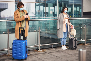 Young man and woman in medical masks using smartphones outdoors