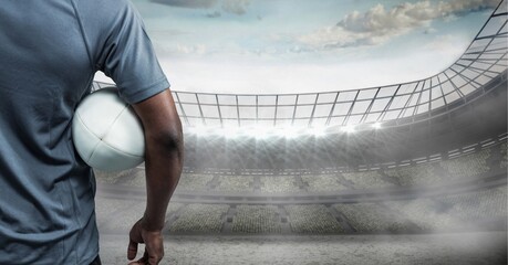Mid section of african american male rugby player holding a rugby ball against stadium in background - Powered by Adobe