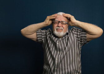 Handsome elderly energetic, charismatic gray-haired man in glasses, holding his head with his hands, emotions in the frame, pain, half-length portrait, blue background