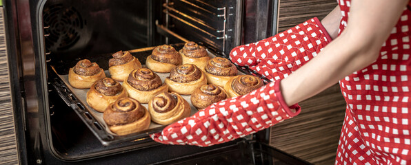 Female hands are taking out fresh fragrant cinnamon rolls from the hot oven. Concept of the...