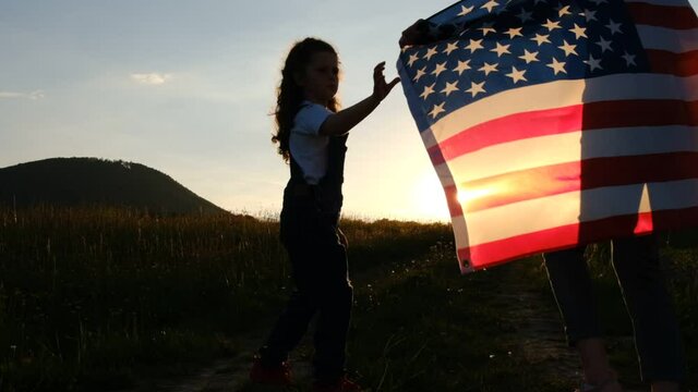 Selective focus of mother with daughter run meadow in wind with united states flag on background mountains during epic sunset. Concept of Memorial Day or 4th of July, Independence Day, Veterans Day