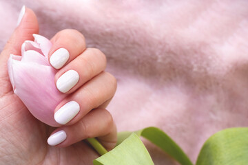 Female hand with beautiful manicure - white ivory nails with tulip flower on blurred pink fabric background with copy space. Selective focus