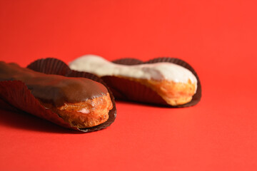 Classic French desserts. Eclairs with white and dark chocolate filled with custard cream served on dessert paper pieces. Two Puffy Eclairs on red background. Choux pastry. Copy space image
