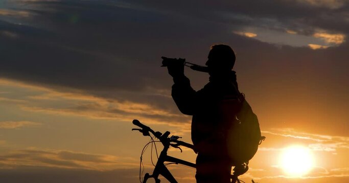 Silhouette of male photographer cyclist stands with camera on road and looks into the distance and admires the beautiful landscape at crimson sunset.  