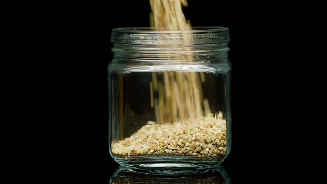 Close-up of falling down green buckwheat into glass jar on black background. Shooting of cereal and groats in studio. Macro footage of pouring out the grits. Food cooking video. 