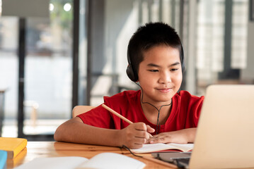 Front view of boy wearing headphones holding pencil taking notes with laptop to study online at...