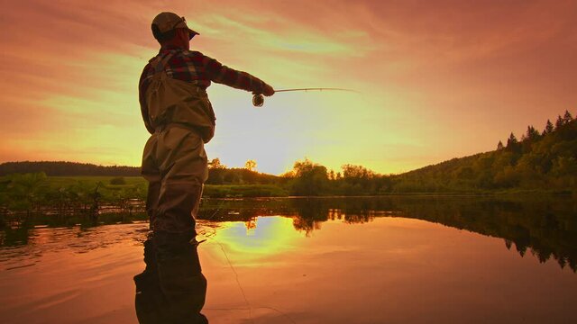 Fly fishing. Angler stands in the river and casts the fly at sunset. Fisherman fishing on fly on the calm river at sunset and casting the line