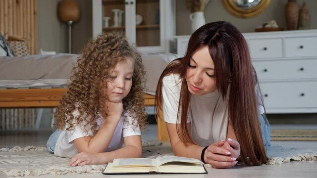 Brunette mother reads book to little daughter with long loose curly hair lying on floor against blurred designed furniture at home close view