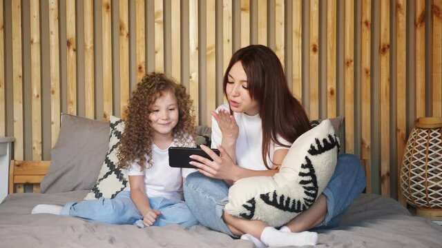 Brunette mother and kid daughter talk with father on video call waving hands and blowing kisses, on large grey bed against brown wooden wall at home