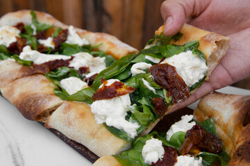 Delicious gourmet food. Italian cuisine. Closeup view of a male caucasian hand holding a slice of fresh arugula pizza with burrata cheese and sun dried tomatoes, on the white marble table. 