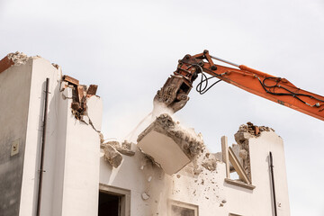 Demolition crusher in action, demolishing an old house, with large chunk of debris falling.