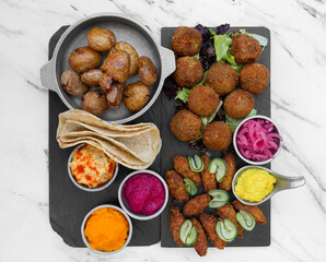 Vegan cuisine. Top view of fried potatoes, vegan fried kibbehs, lentil falafels, onion pickles, garbanzo, beets and carrot hummus, and corn tortillas on the white marble table. 