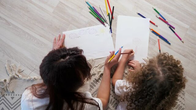 Brunette mother with long hair helps little girl draw picture on sheets of papers with coloured pencils lying on brown floor close upper view