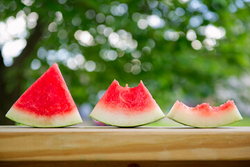 Fresh slices of watermelon on a wooden table in front of a blurred background featuring tree leaves.