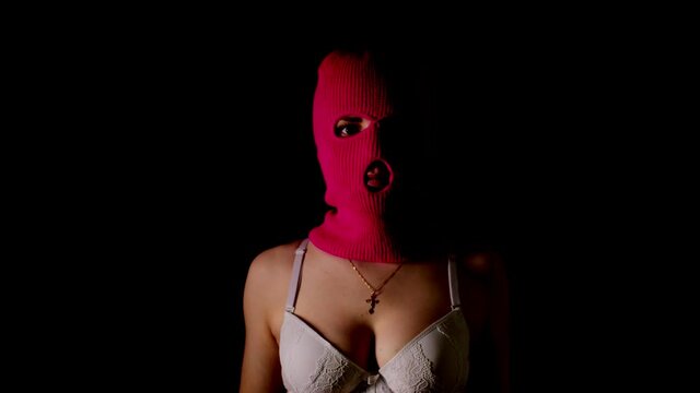 Unrecognizable woman in pink balaclava and white bra stands in dark. Unknown female in mask and lingerie highlighted in dark room.