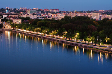 Fototapeta premium Ternopil, Ukraine-May, 11,2021:Night lights landscape of Ternopil. Motorway between city lake and park with green trees. Colorful light trails with motion blur effect. Romantic and peaceful scene