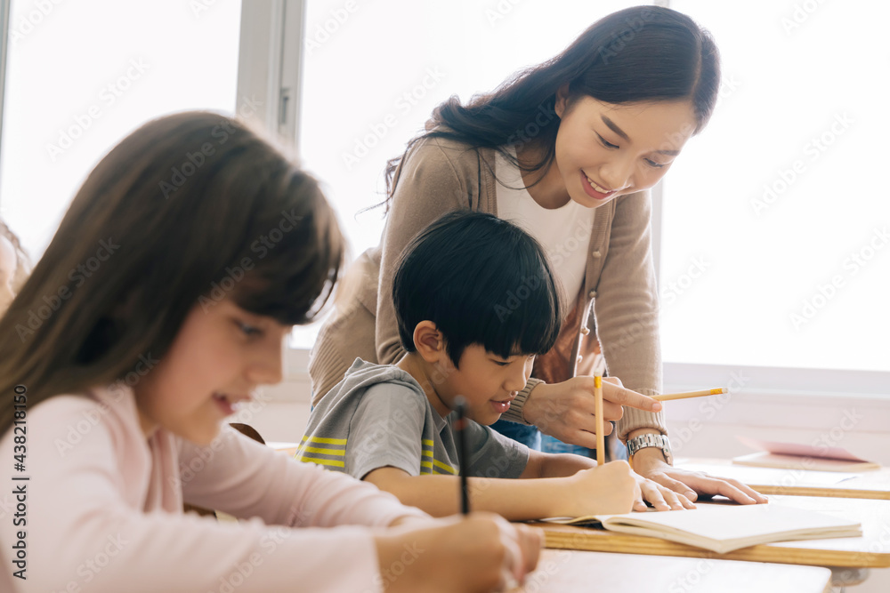 Wall mural asian school teacher assisting students in classroom. young woman working in school helping boy with