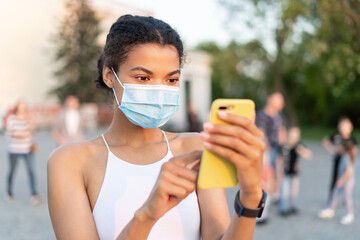 Young African American woman wearing face mask outside and using her mobile phone during...