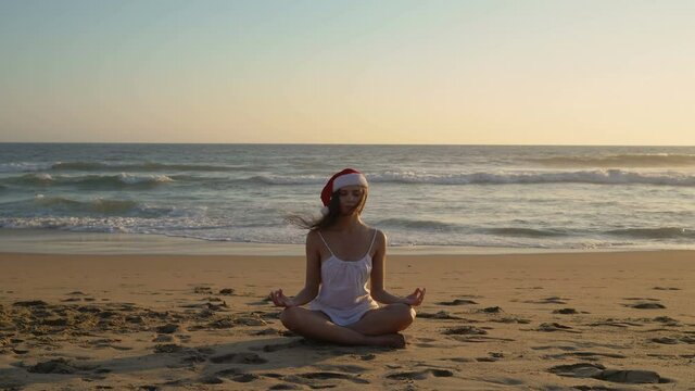 Attractive young woman in a white dress and a red Santa Claus hat. Exotic exterior The girl meditates on the coast of the Pacific Ocean.