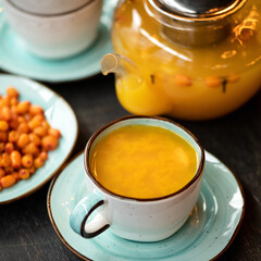 Full Cup of orange sea buckthorn tea, close-up shot. Vitamin healthy drink useful for strengthening immune system. Fresh berries and glass teapot with healing drink on blurred background. Soft focus.