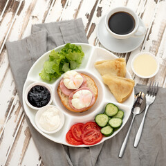 Traditional European or American Continental breakfast on rustic wooden table with linen tablecloth. Thin pancakes, sausage, bun, poached eggs, salad, sauces and cup of coffee on portioned plates.