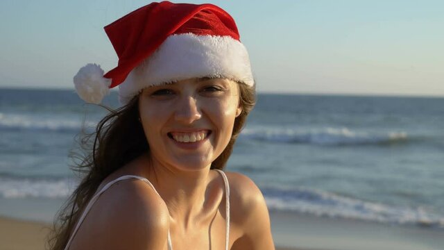 Winter holidays in a warm country. Young adult woman in Santa Claust Hat stands on the beach. Portrait of a long-haired woman.