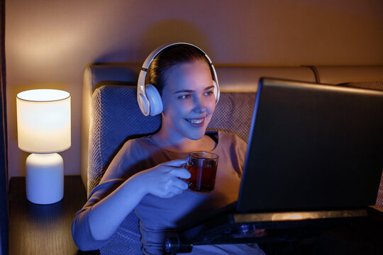 Smiling teen girl in headphones watching video on laptop in the evening alone. Young happy woman watching movie using laptop and drinking tea at night while lying on couch.
