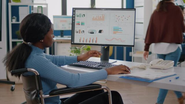African employee with disabilities, handicapped paralyzed woman checking graphics from computer working in financial start up business office. Busy focused project manager typing on pc in workspace