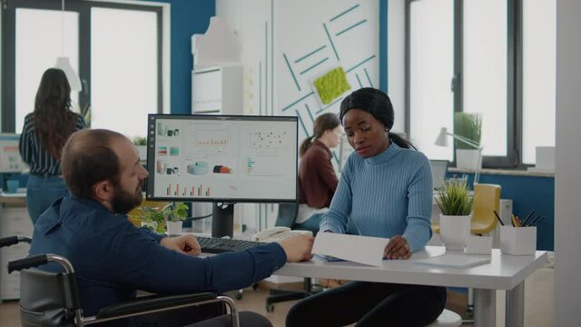 Black manager woman working with financial documents checking graphs talking with paralysed team leader with disabilities, reading raports sitting in wheelchair in start-up business office