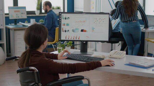 Handicapped businesswoman analysing financial statistics sitting in wheelchair checking graphs from computer in start up business office. Disabled employee working with team using modern technology