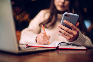 Fototapeta premium Crop female manager taking notes in daily planner while messaging on phone in cafe