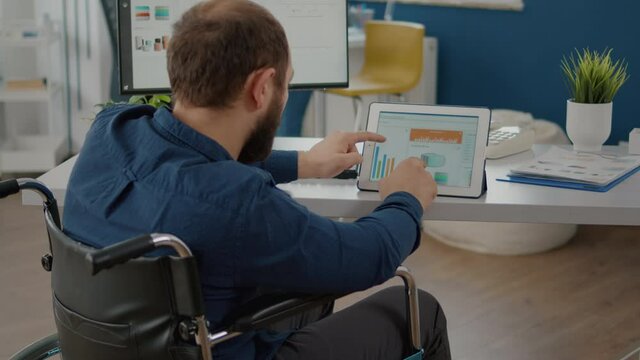 Disabled handicapped immobilized paralyzed manager man using computer and tablet in same time working in business start-up office sitting in wheelchair. Busy multitasking employee typing searching
