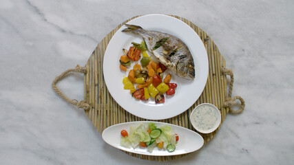 Serving roasted sea bream fish with vegetables, sauce, and fresh salad. Top view