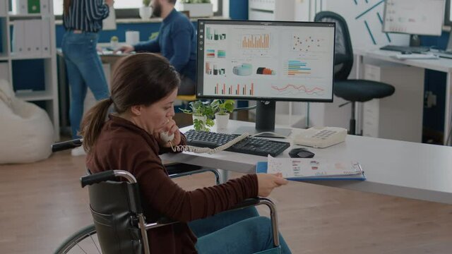 Busy paralysed handicapped woman worker talking at phone with business partner sitting in wheelchair in start up company office resolving financial problems. Teamworkers discussing statistics graphs