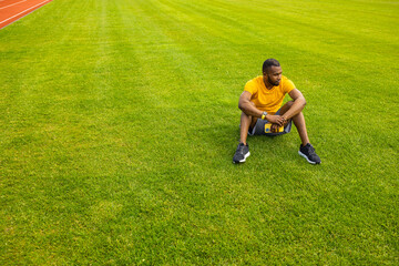 Fototapeta na wymiar Young pensive Afro American sportive male sitting with smartphone on grass in the middle of stadium, relaxing after workout training, football soccer match. Copy space. Healthy sport lifestyle concept
