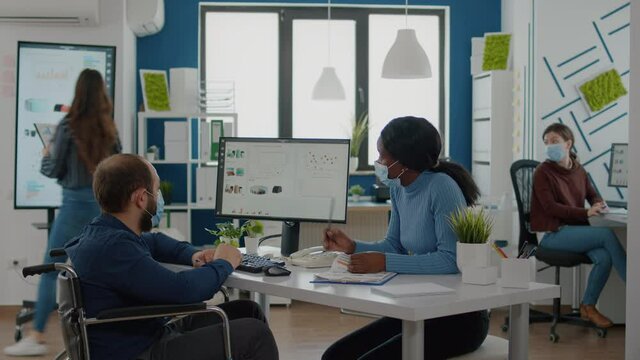 Paralysed freelancer with face mask discussing with black woman partner financial problems, checking statistics informations sitting in new normal start up office pointing at computer analysing graphs