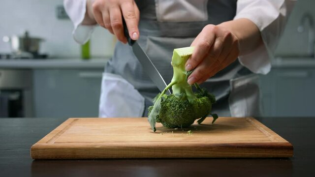 Female hands separates broccoli florets from stem by knife on kitchen background
