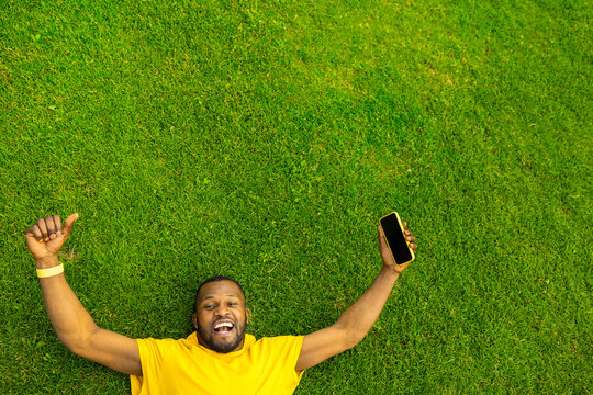 Top view of cheerful overjoyed African American male celebrating victory, successful race, game or football match. Young afro laying on the grass field stadium, holding mobile phone and show thumb up