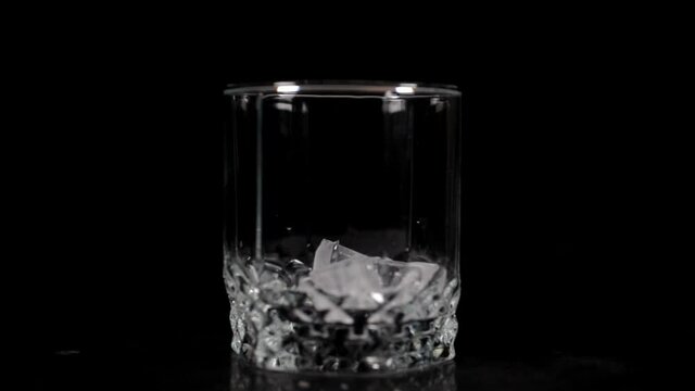 Bartender throw ice cubes into a rotating glass on a black background and pours whiskey into it, close-up, selective focus