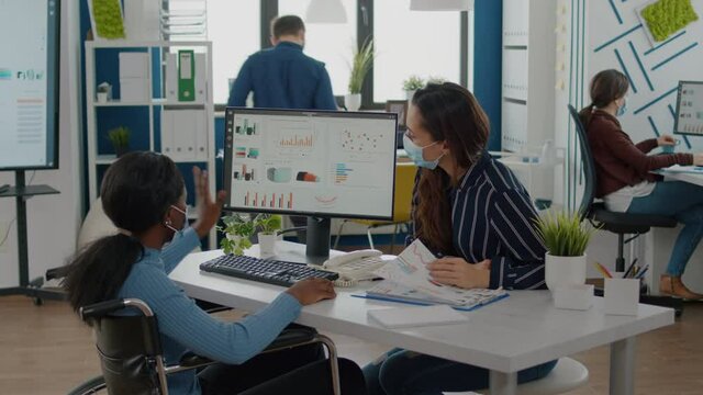 Immobilized handicapped invalid african entrepreneur working with colleague in new normal business office during covid19 pandemic wearing face masks, checking graphs calculating profit on computer