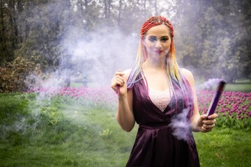 A girl with a smile with bright makeup and colored braids in a purple dress blows artificial purple smoke in a spring blooming park