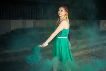 Obraz premium Cute girl in a green dress with African braids and colorful makeup posing with bright green smoke on the streets of a warm spring city