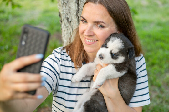 Young beautiful woman in a t-shirt and jeans with a husky puppy takes a photo or selfie on the phone while sitting in a park near a tree