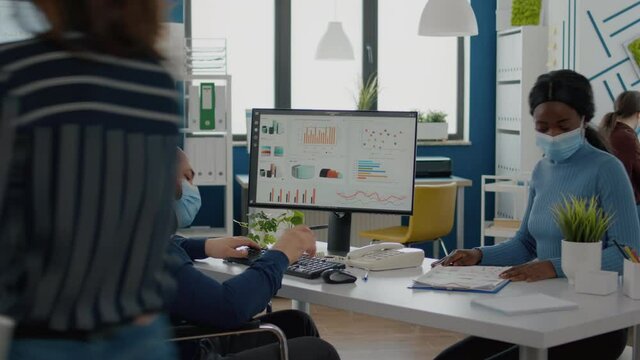 Diverse colleagues with masks respecting social distance working with financial graphs in new normal stratup office, black woman and paralysed coworker in wheelchair discussing solution for project