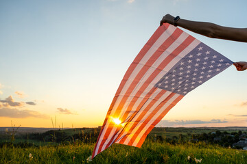 Human hands holding waving USA national flag in field at sunset.