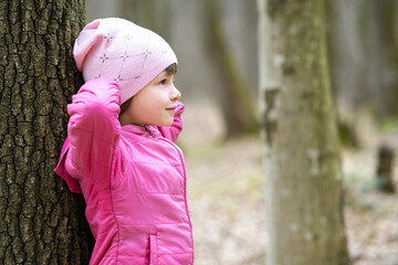 Portrait of young pretty child girl wearing pink jacket and cap leaning to a tree in forest...