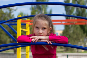 A child girl with blond hair in a pink T-shirt sits pensive and sad on the playground. 